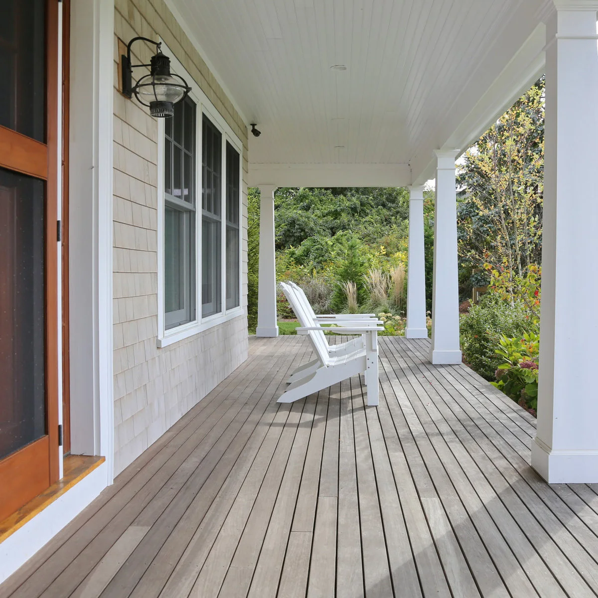 A long porch beside pale shingle siding has white square columns, a white ceiling, wood floorboards and two white Adirondack chairs.