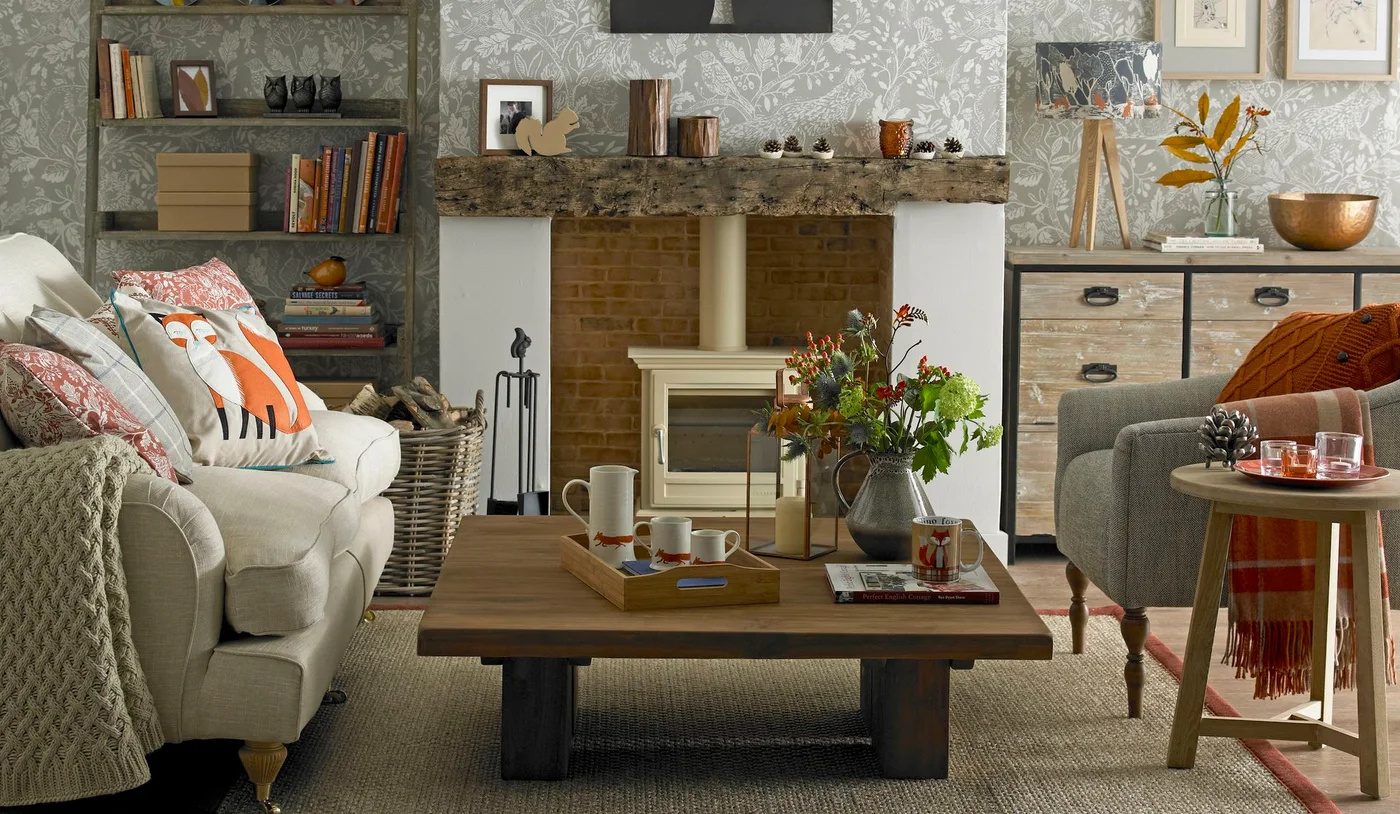 A rough wooden mantel sits above a pale stove beside woodland-patterned walls, a white sofa and a wooden table
