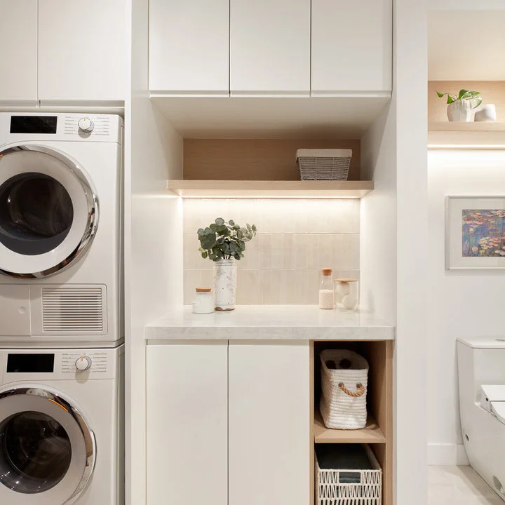 Stacked white machines sit beside plain white cabinets, a counter and a light wood niche near a bathroom doorway.