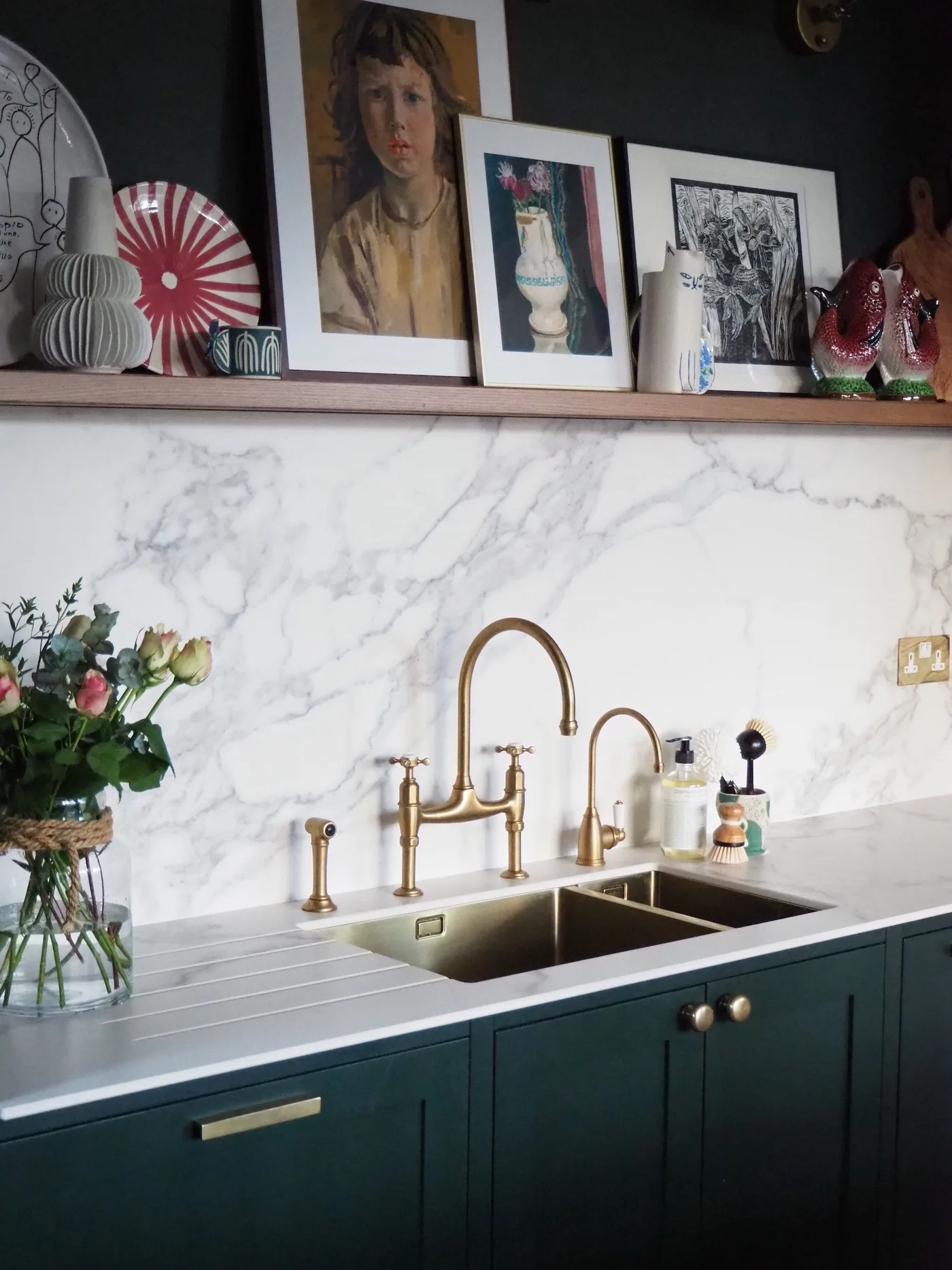 Dark green cabinets sit beneath a double sink and pale veined backsplash with a wooden shelf of framed pictures and decorative plates