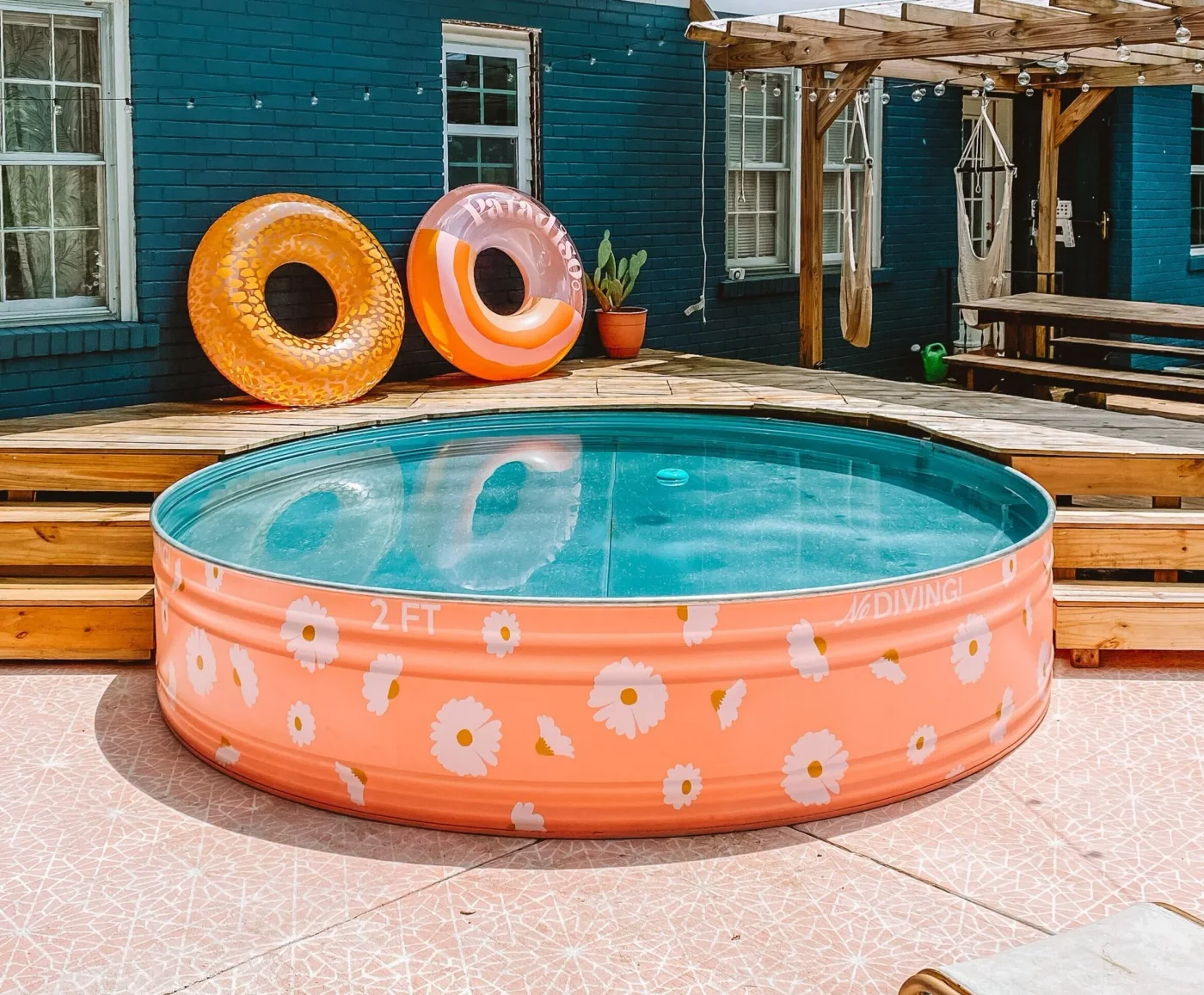 Peach-colored stock tank pool with white daisy shapes in front of a teal wall and wood deck.