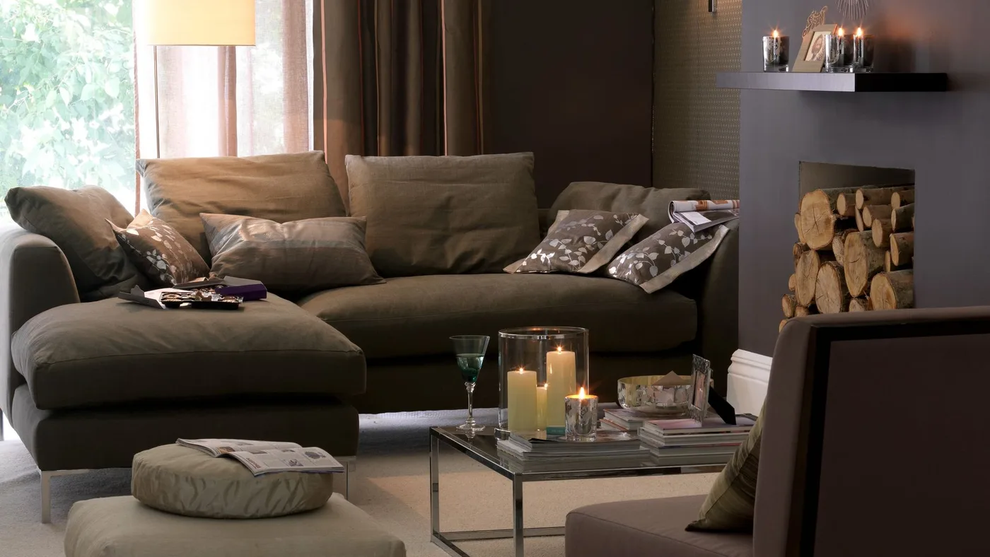 Brown fabric chaise sofa with floral cushions, a glass coffee table and brown walls.