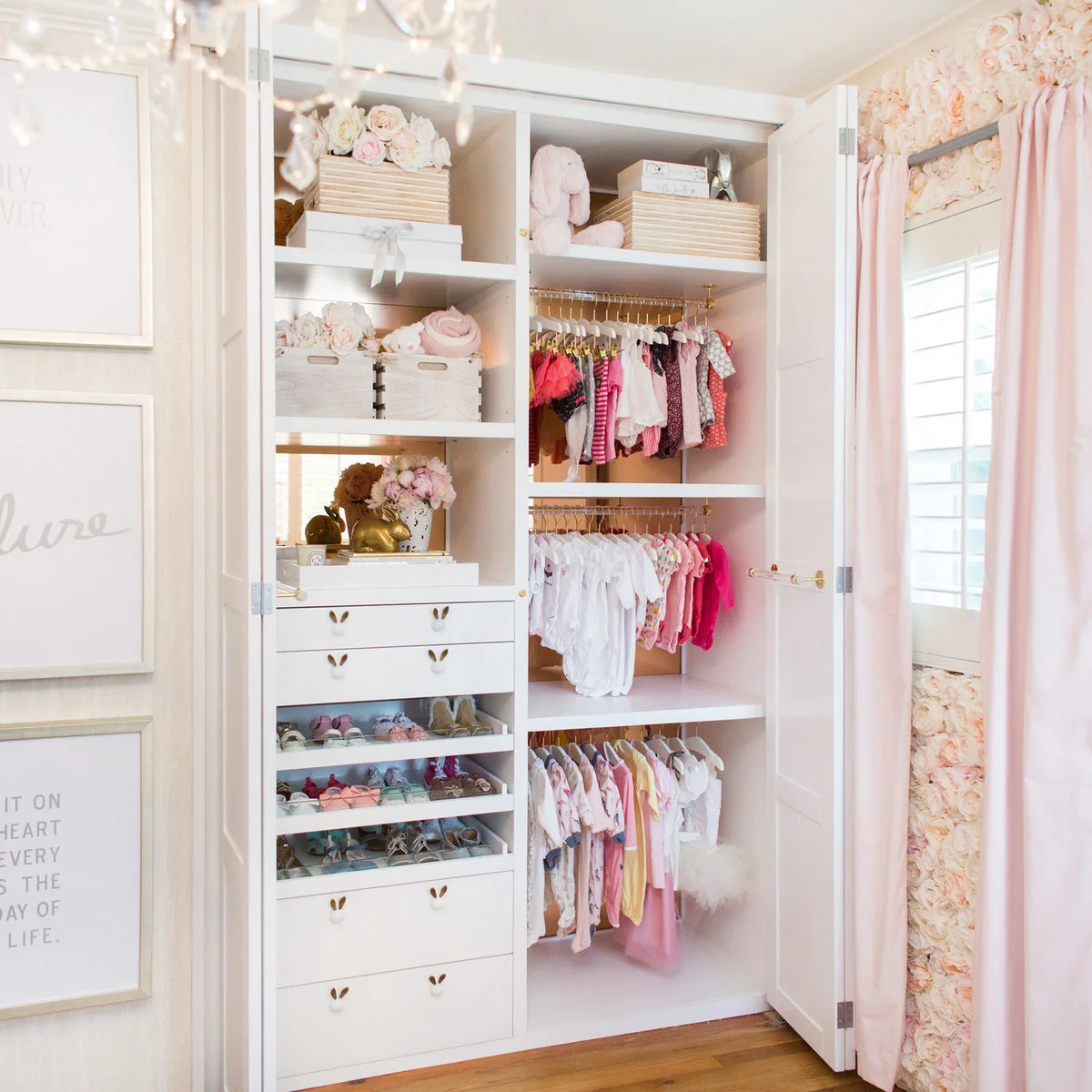 Open nursery closet with several short hanging rails, white drawers, shoe shelves and storage baskets