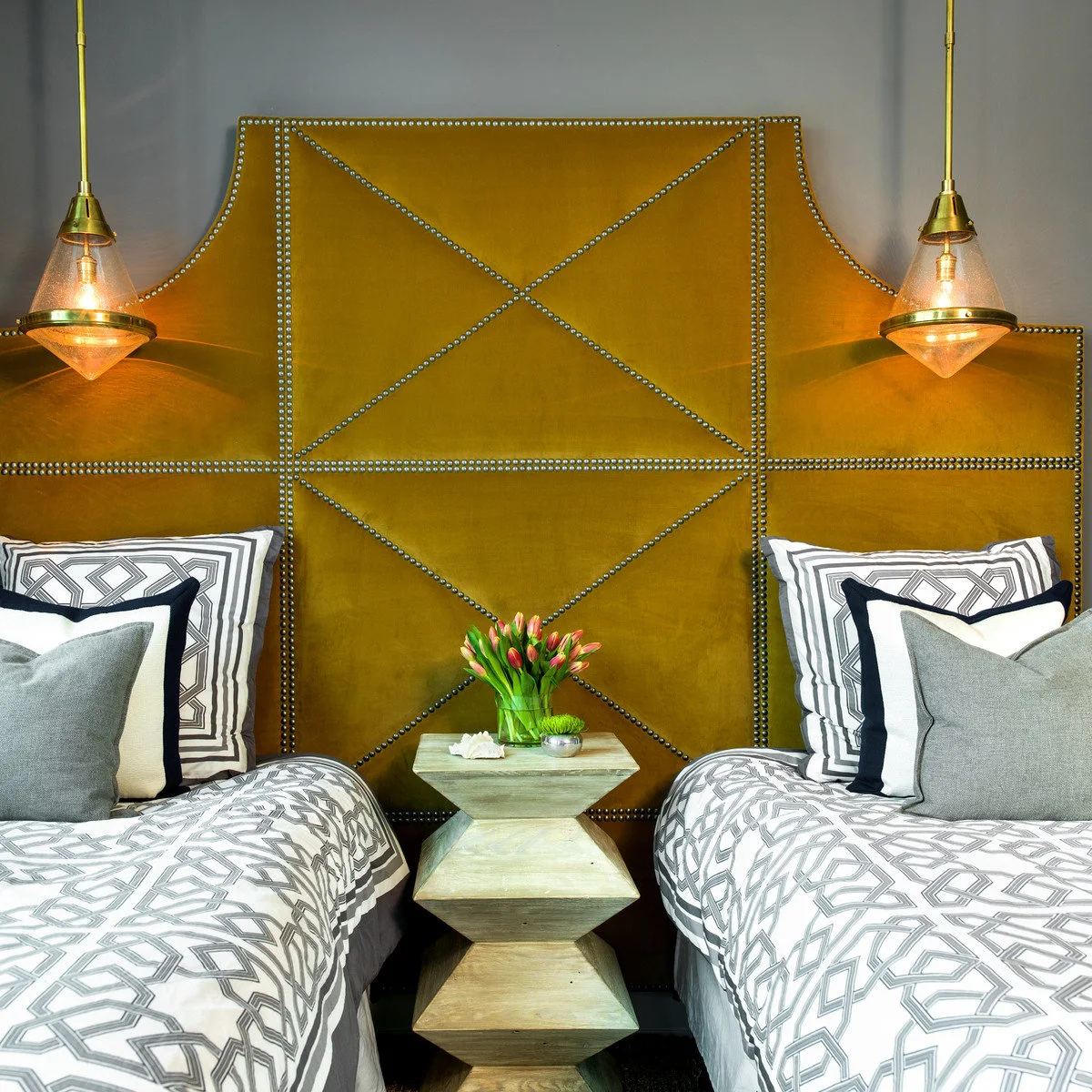 Two beds with gray patterned covers and a narrow sculptural table against a mustard upholstered headboard