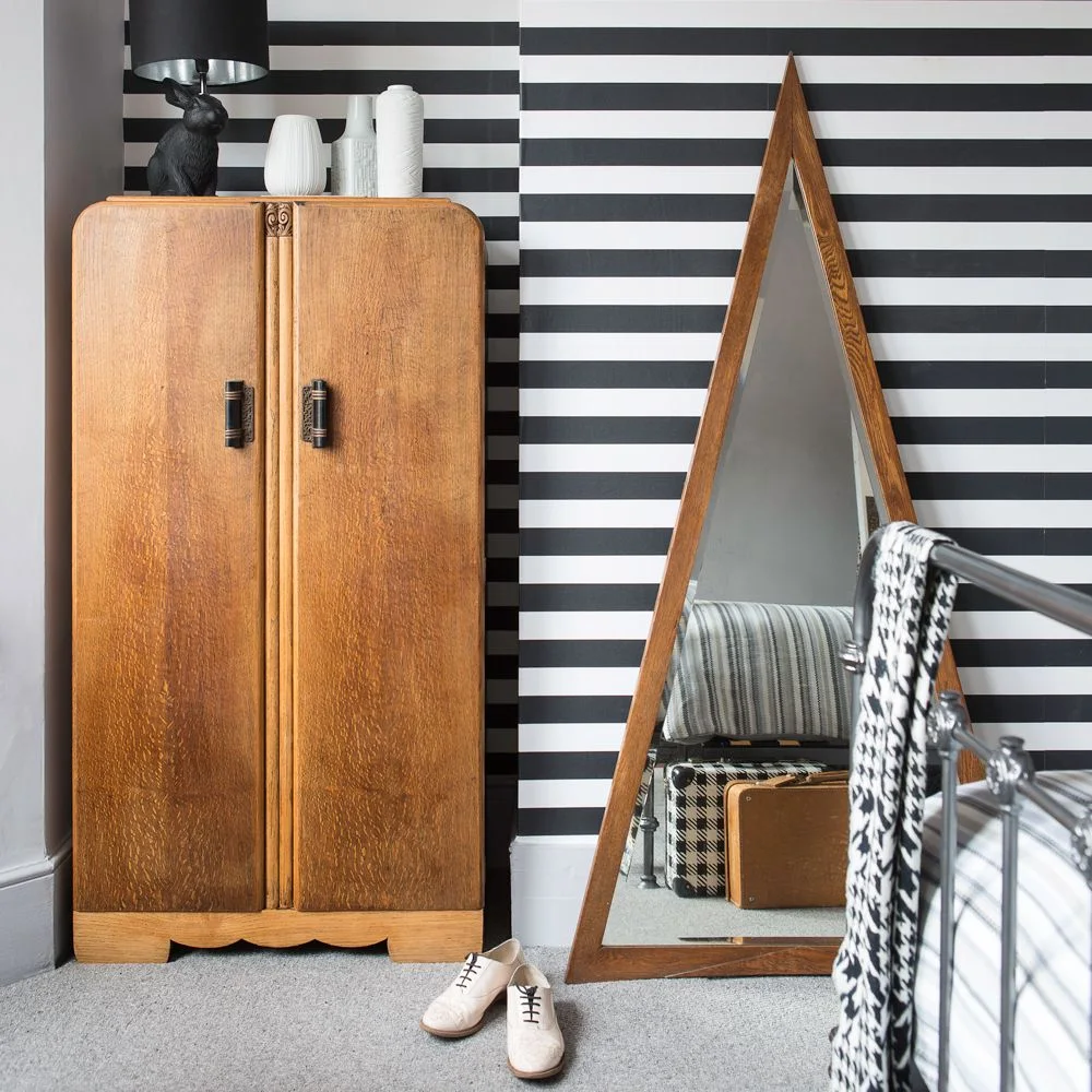 Black-and-white horizontal wallpaper stands behind a warm wood wardrobe and tall triangular mirror, beside a metal bed and gray carpet.