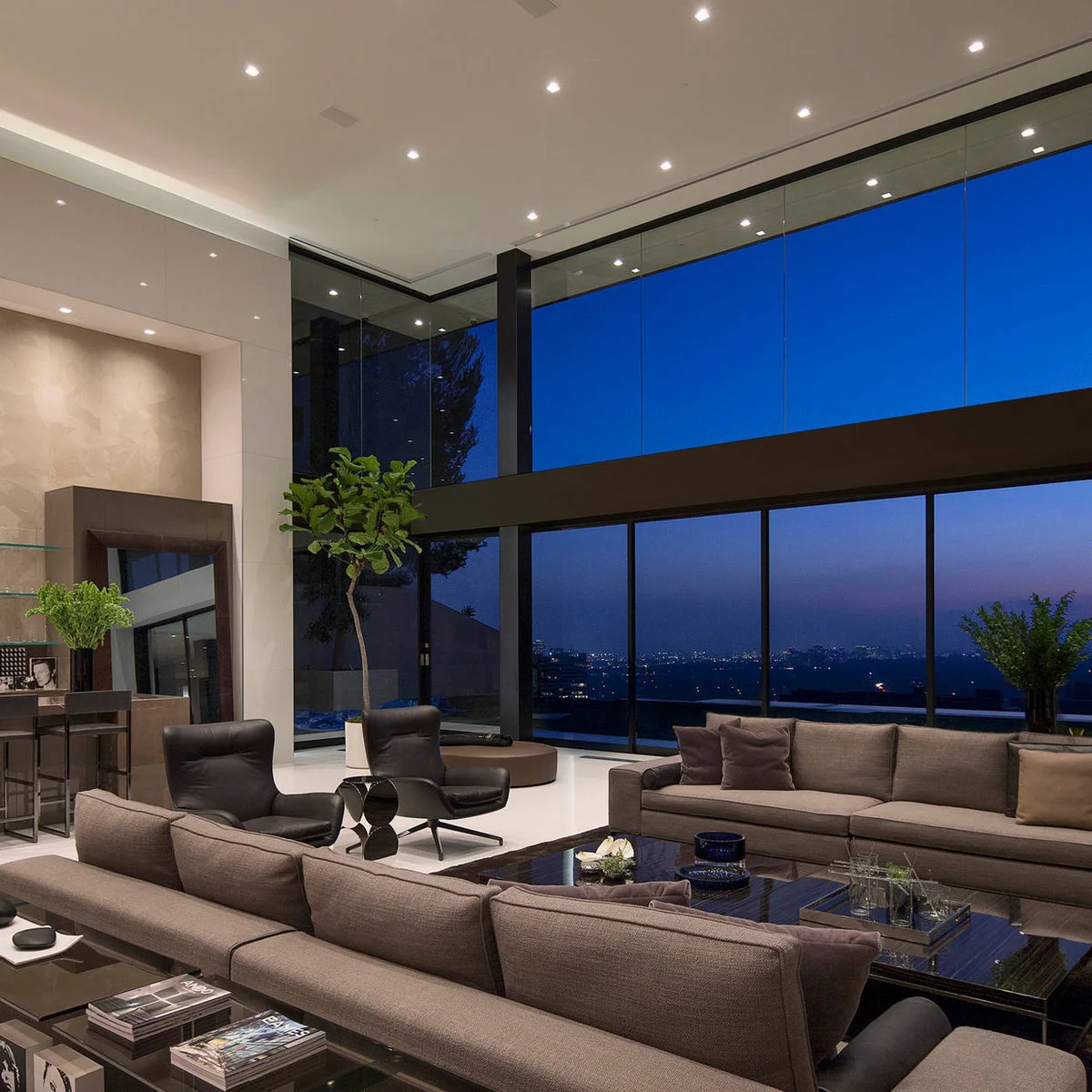 A tall living room with two tiers of dark-framed glass, gray sofas and black lounge chairs around glossy coffee tables.