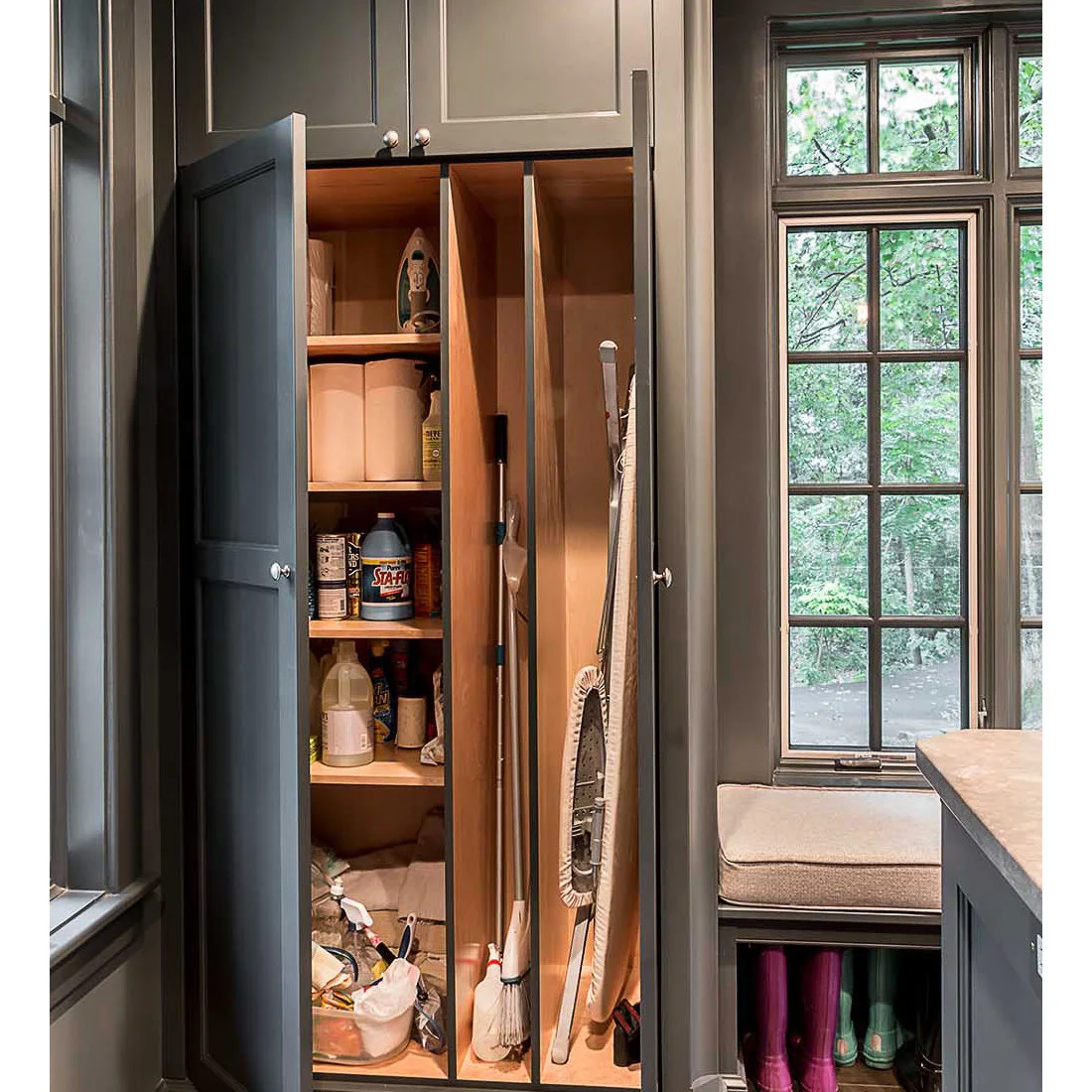 Open dark laundry utility cabinet with shelves, a narrow broom-and-ironing-board bay and a window seat beside it