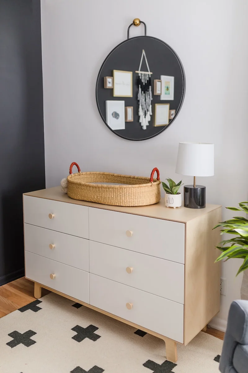 A pale wood-toned nursery dresser holds a woven basket below a dark round mirror beside a white-shaded lamp and pale rug with dark crosses