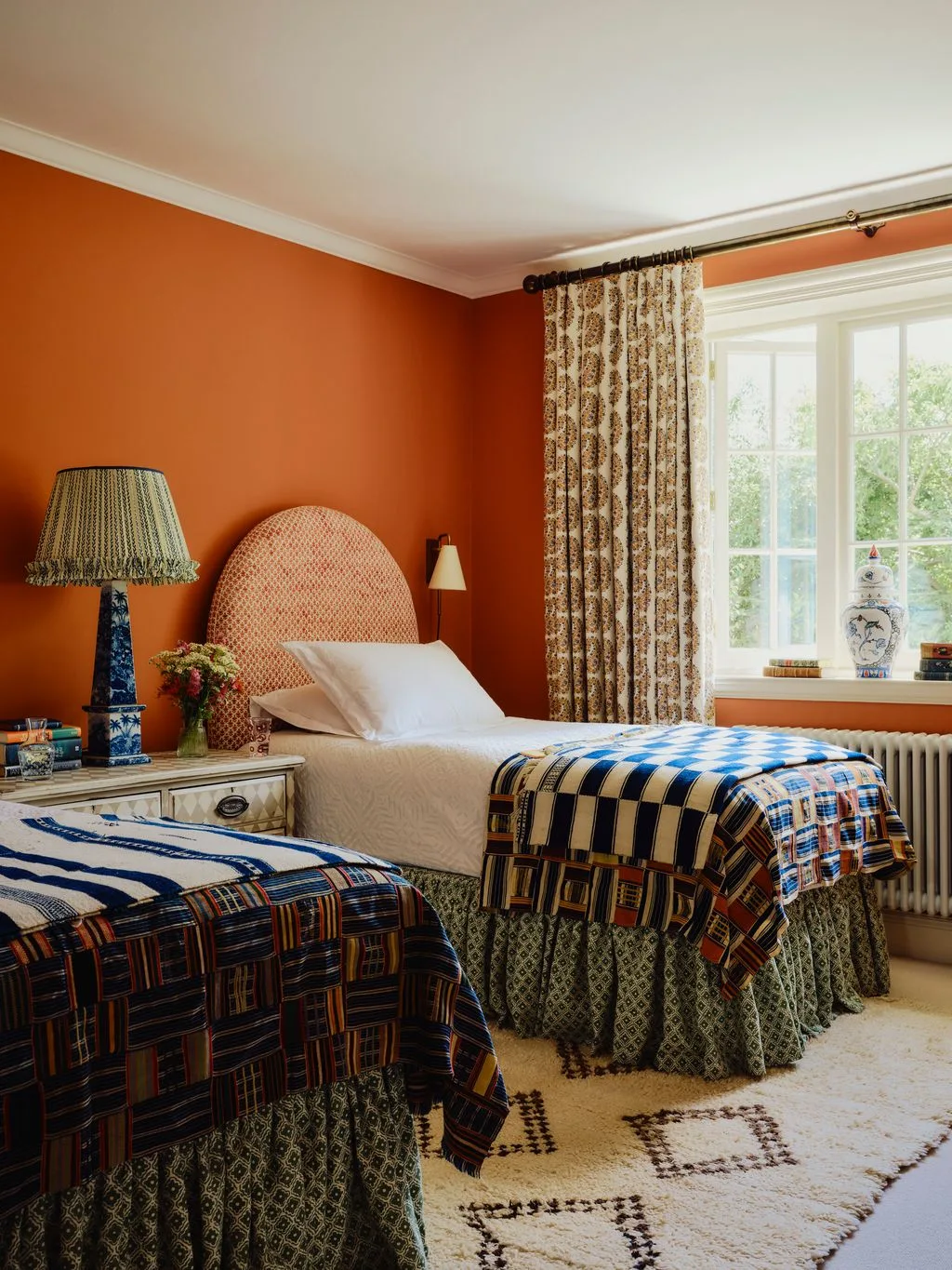 Twin bedroom with orange walls, patterned curtains, rounded pale headboards, blue checked throws and a tall blue bedside lamp