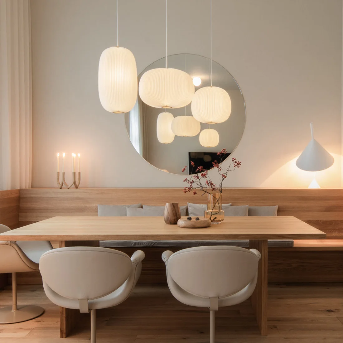 Three rounded white hanging lights sit above a wood dining table, pale chairs and a long cushioned wood bench beneath a round mirror.