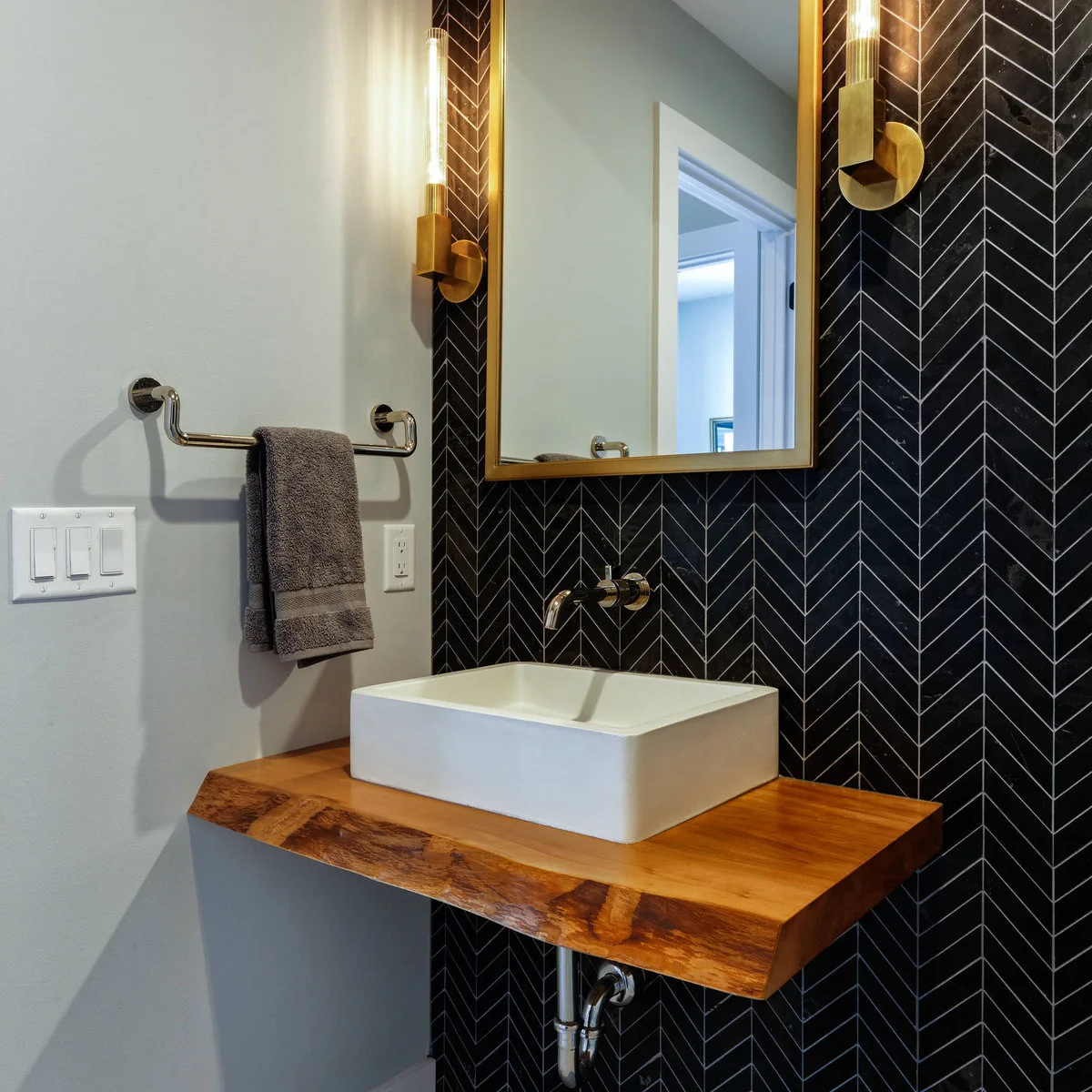 Two warm metal wall lights with tall clear lamps flank a warm metal mirror above a white vessel sink and dark chevron tile.
