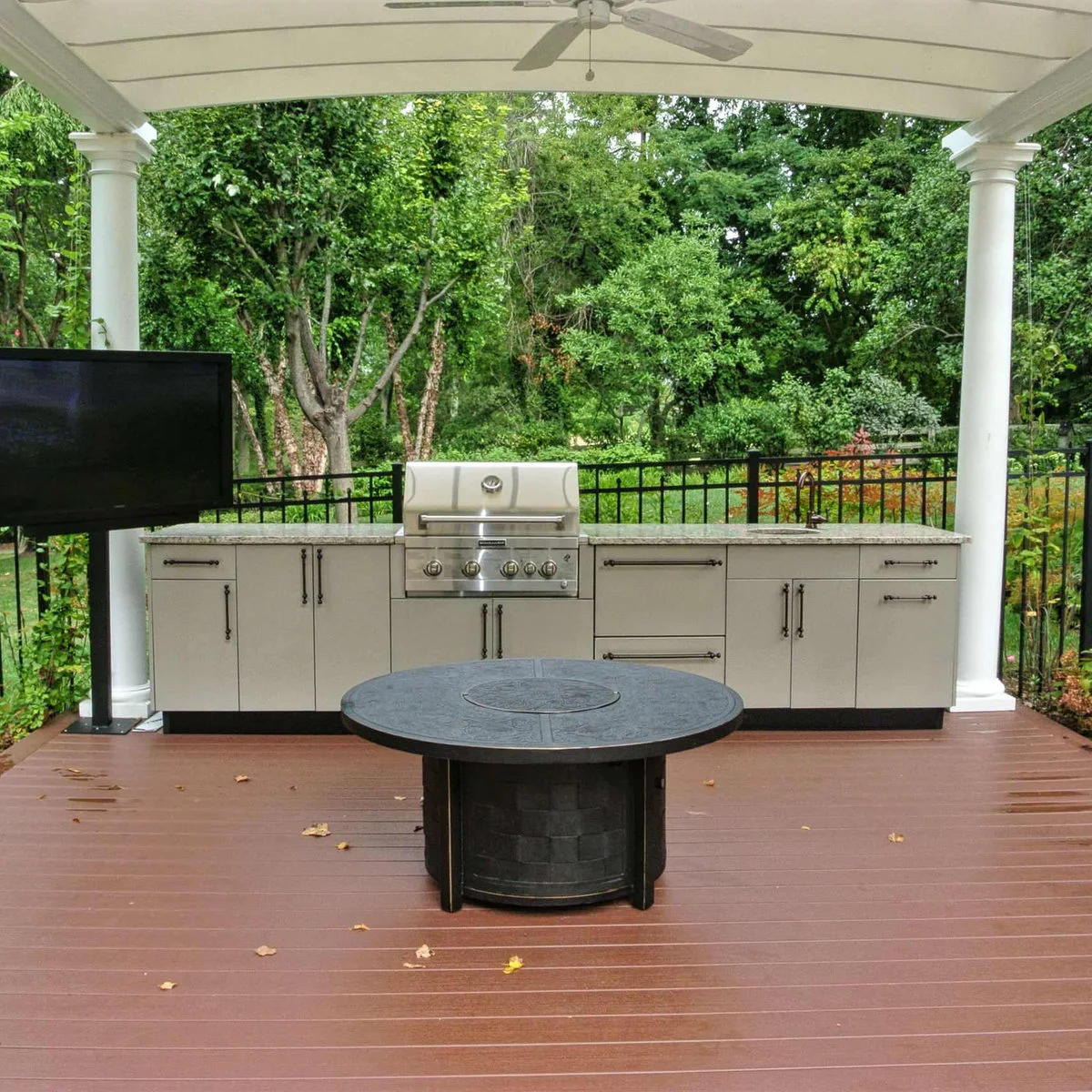 Straight outdoor kitchen with pale cabinets, central grill, small sink, black railing and a round table in front
