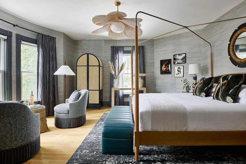 An open wood canopy bed with white bedding and dark leaf-patterned cushions stands beside gray chairs, a teal bench and a woven-inset arched cabinet