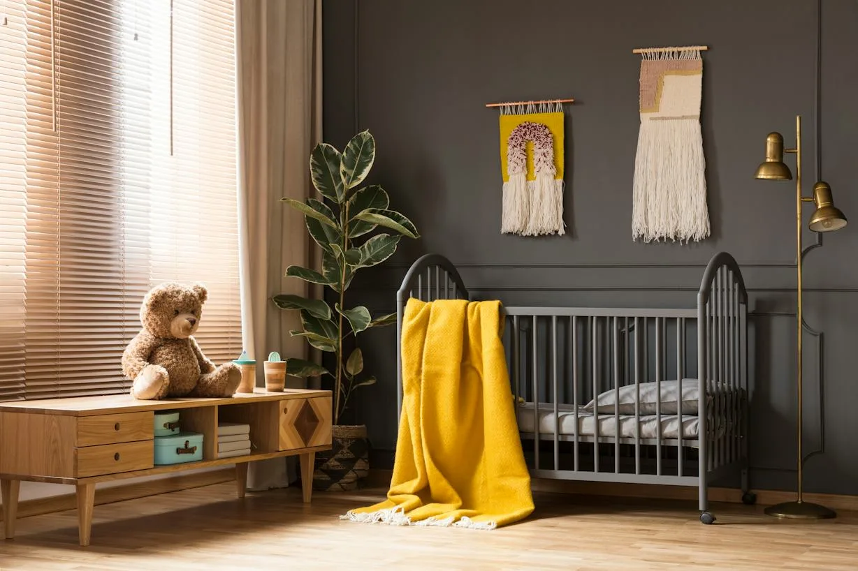 Gray crib against darker paneled wall beside warm wooden storage, mustard staged throw, fringed hangings, plant and floor lamp.