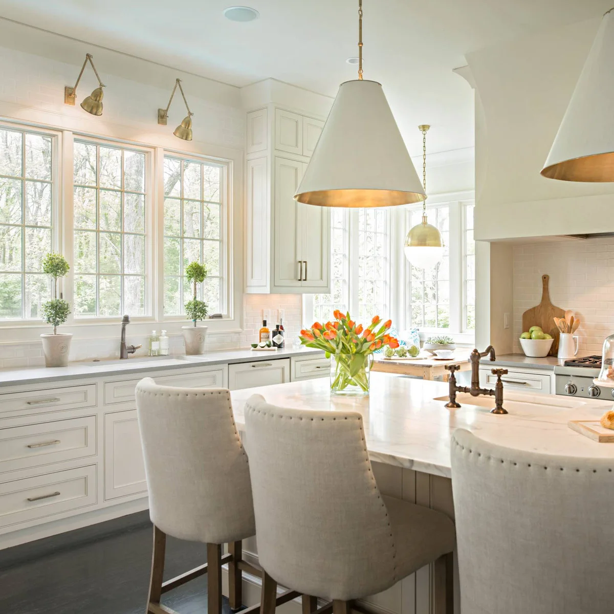 Small warm metal articulated wall lights sit above a broad kitchen window behind large pale cone pendants with warm interiors.