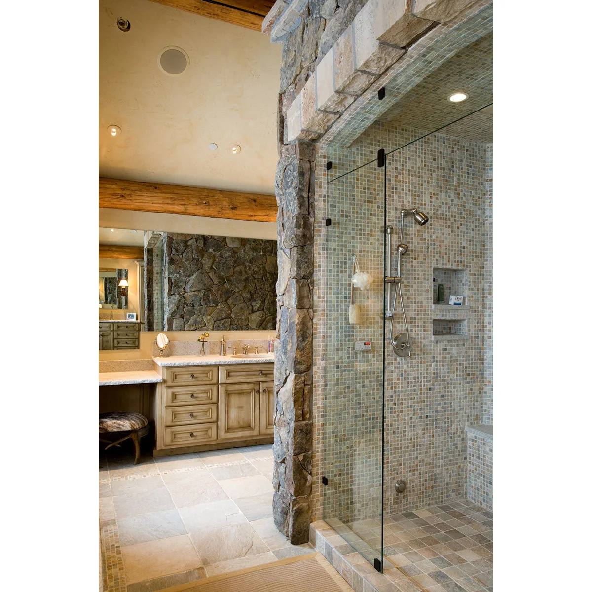 A mosaic-tiled shower behind glass with a broad rough masonry surround and recessed product shelves.