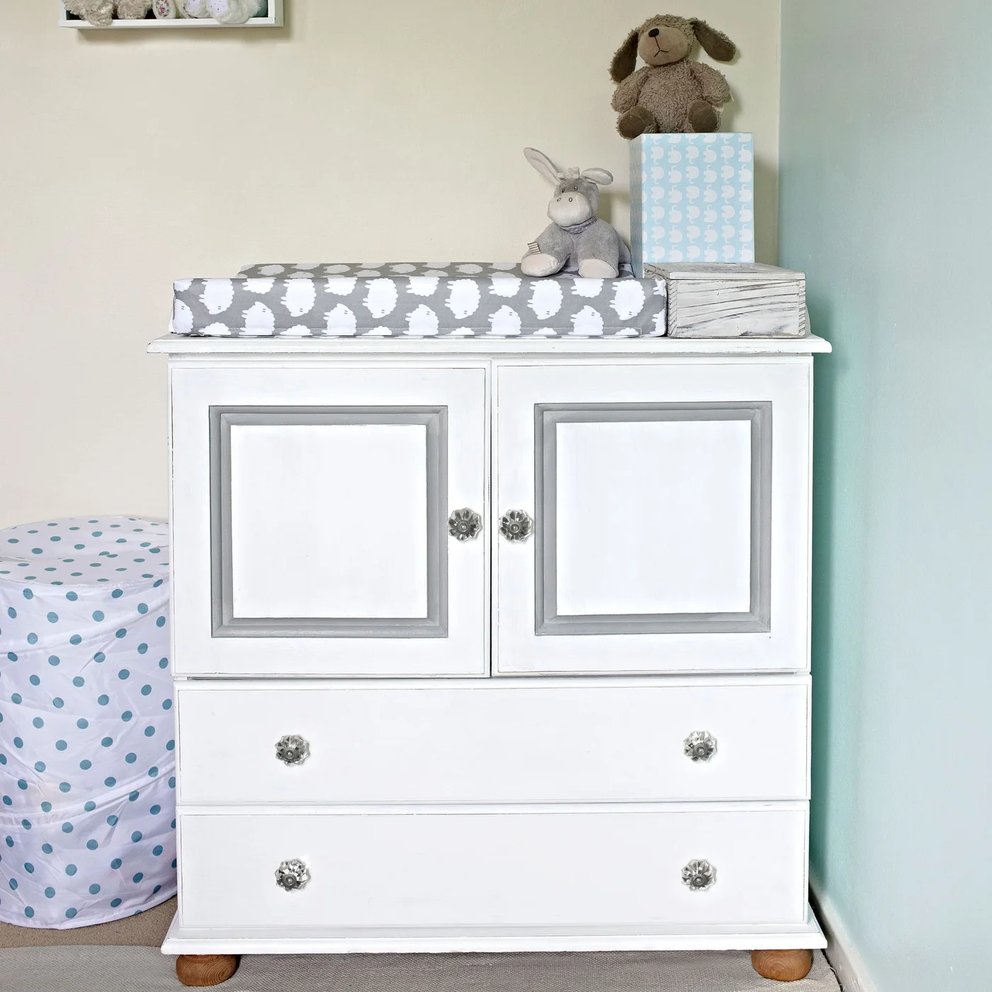 A white nursery cabinet with gray inset trim has upper doors, two lower drawers and a changing pad on top.