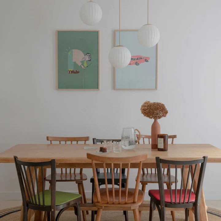 A plain wood table with mixed spindle-back chairs and colored seat pads stands beneath three white globe pendants.