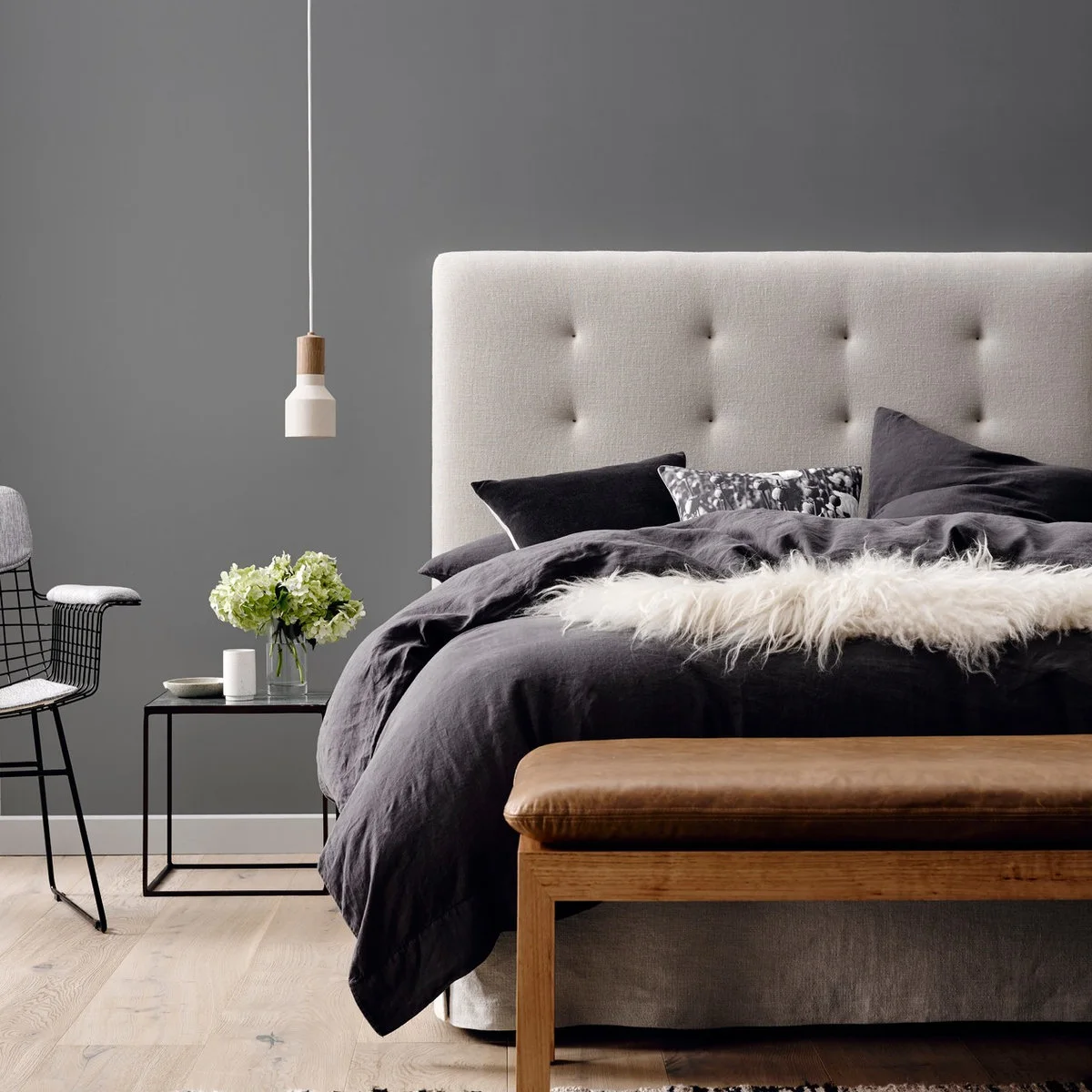 Grey bedroom wall behind a pale tufted headboard, dark bedding and a wood bench