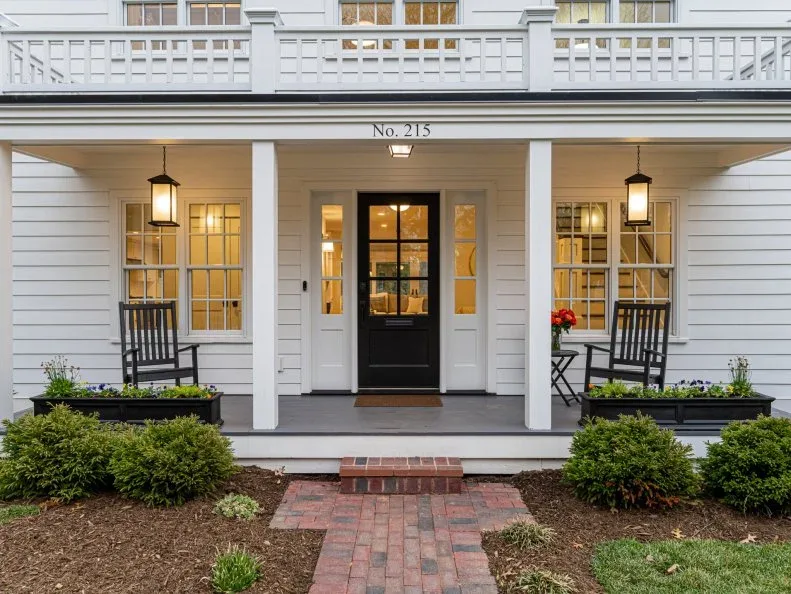 White farmhouse-style porch with black glazed door, dark rocking chairs, lanterns and brick path