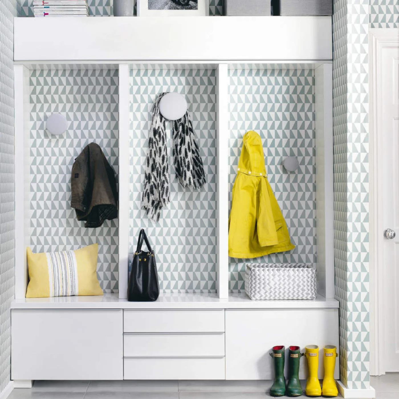 White flat-front cupboards and drawers sit below three wallpapered coat bays with round hooks, with a long upper cabinet above.