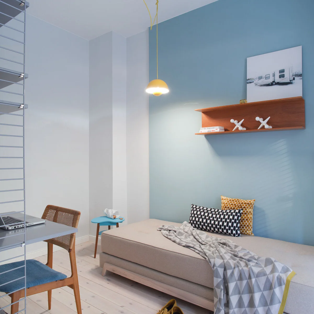 Pale blue wall behind a neutral daybed with patterned pillows, a wooden shelf and a yellow pendant.