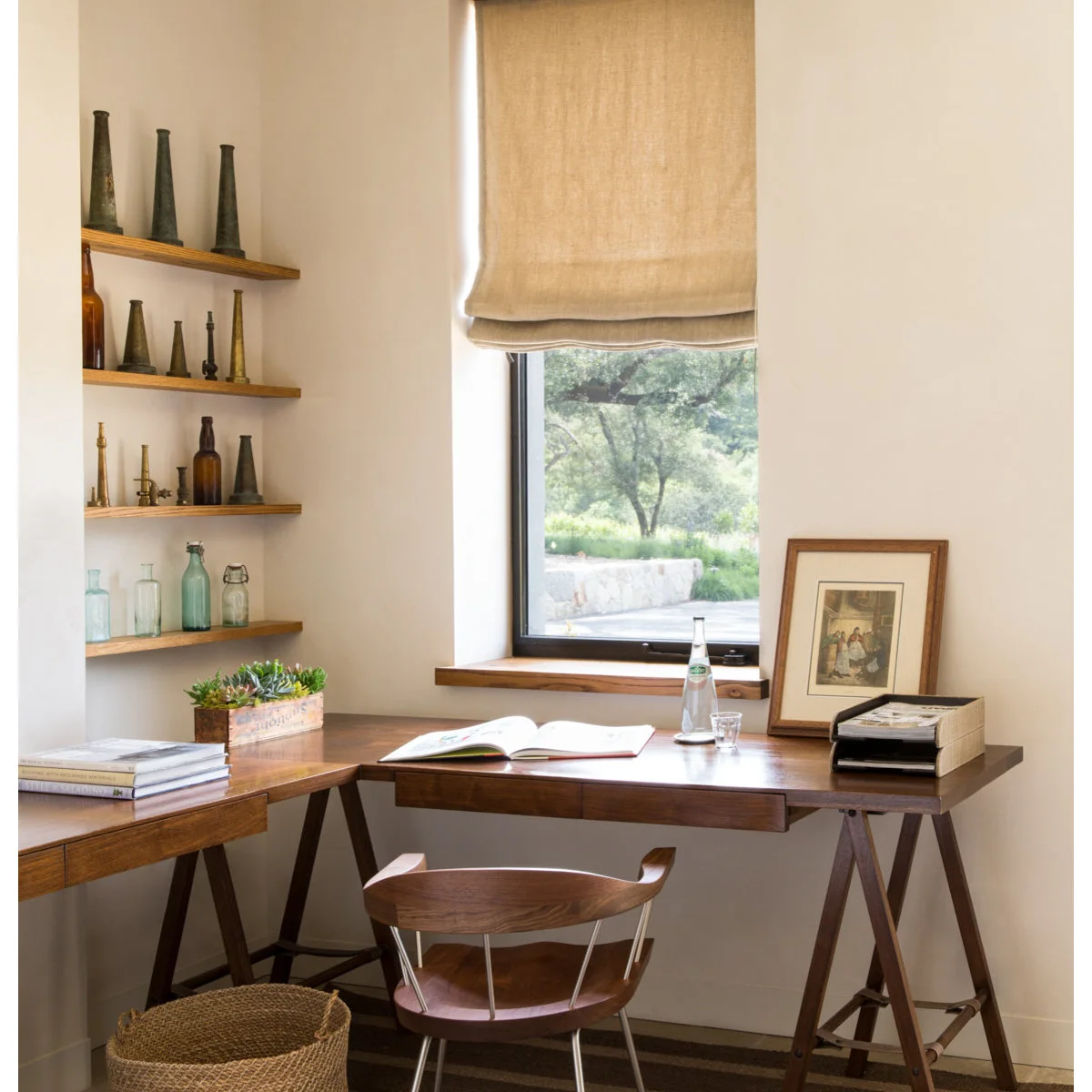 Home office corner with wood trestle desks, woven-seat chair, pale walls and a fabric window shade