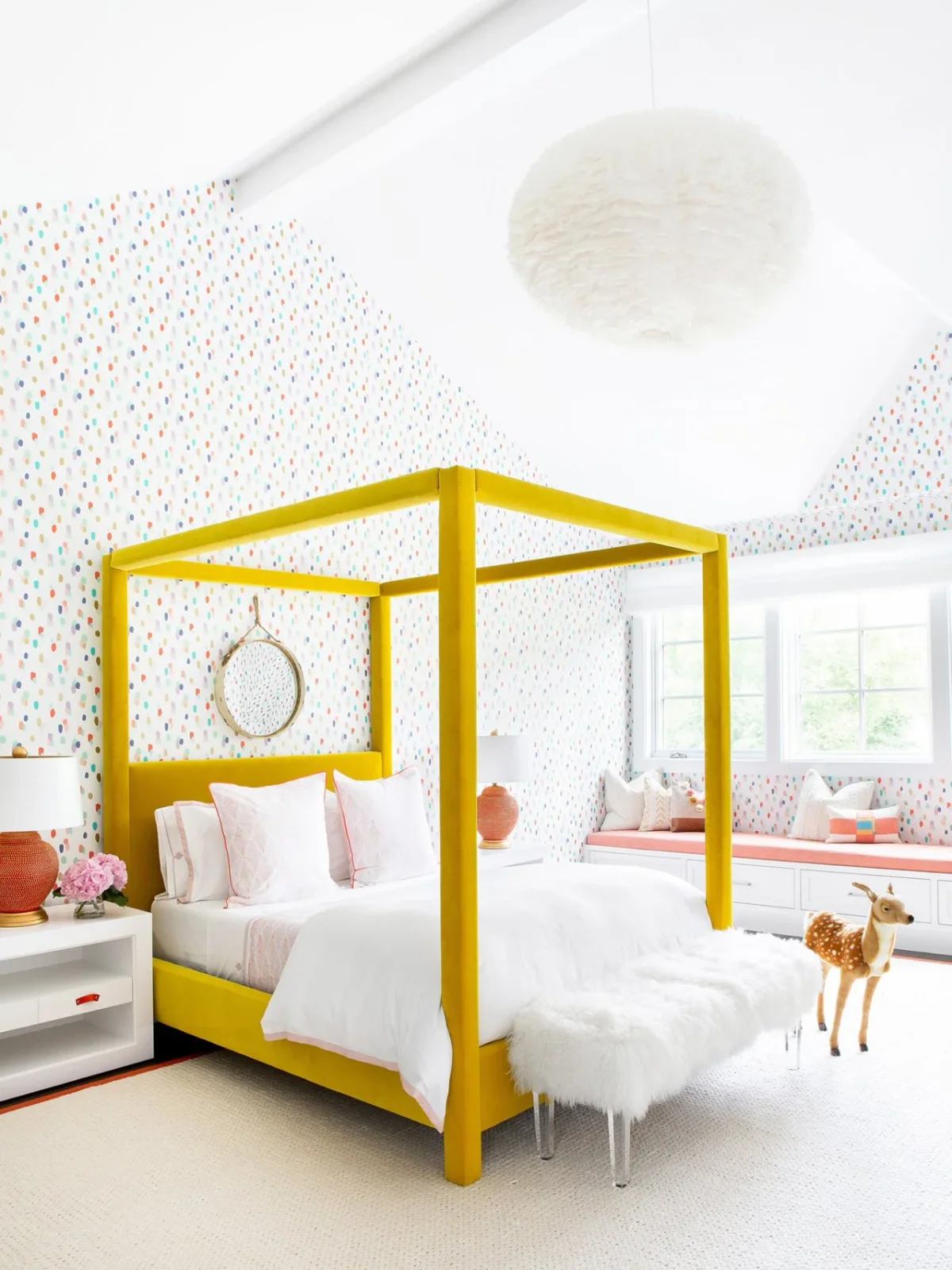 A bright yellow upholstered four-poster bed stands in a pale dotted bedroom with white bedding, pink edging and a peach-cushioned window seat.