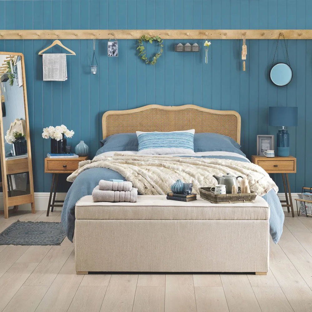 Bedroom with blue vertical paneling, a woven headboard, wooden bedside drawers, pale upholstered bench and a round hanging mirror.