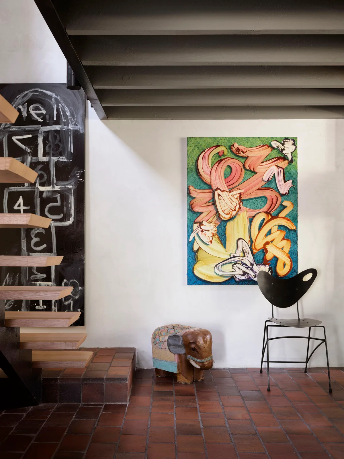 Bright abstract artwork hangs beside a black sculptural chair and elephant-shaped stool near floating wooden stairs above reddish floor tiles