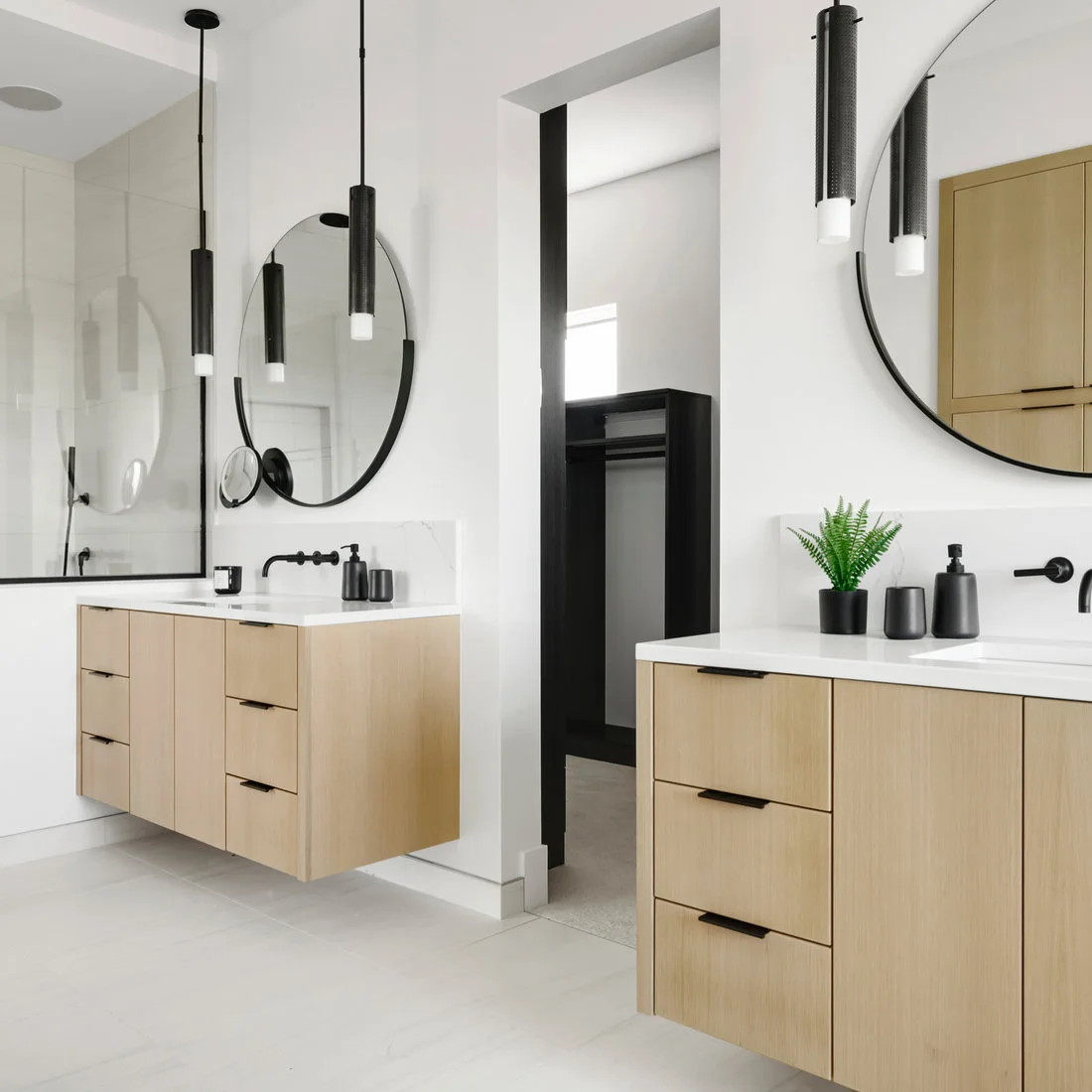 Two floating light wood vanities with round mirrors and black pendants stand on either side of a doorway.