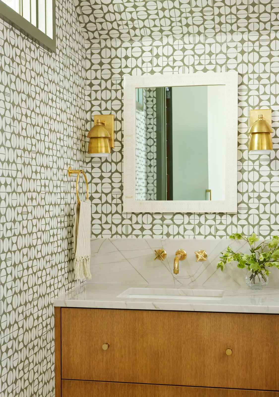 Green and white geometric bathroom wallpaper surrounds a white-framed mirror and gold-colored lights above a wood vanity and pale tiled backsplash