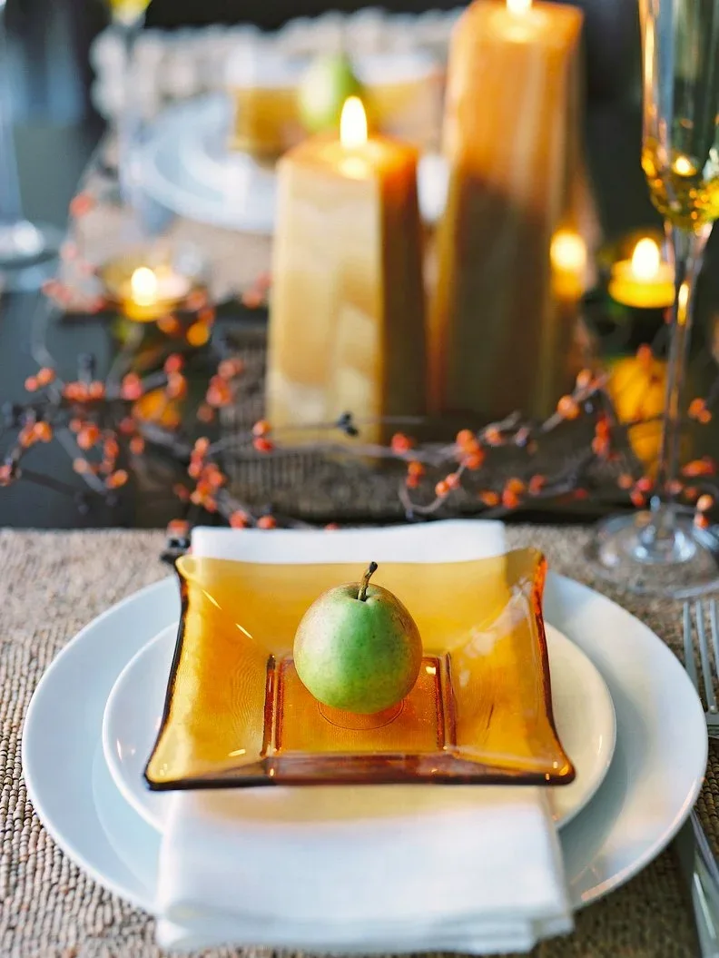 A green pear rests on a square amber-colored plate above white crockery beside pillar candles and a textured pale mat