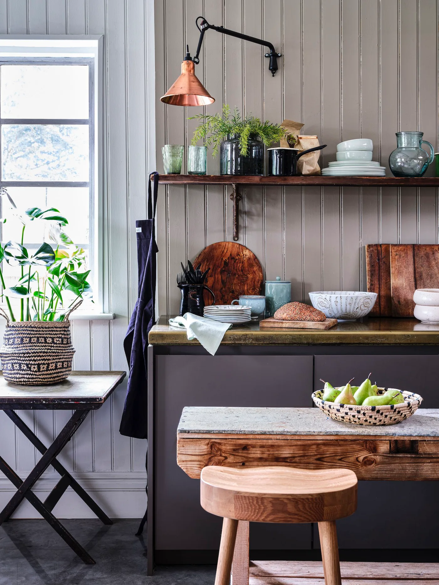 Kitchen corner with gray paneled wall, wooden shelf, copper-colored wall light, dishes and compact wooden surface.