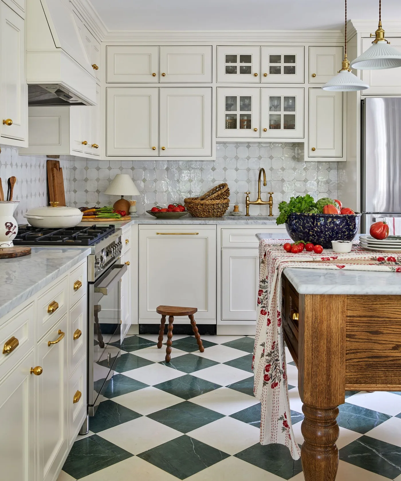 Cottage kitchen with pale framed cabinets, green-and-white checked floor, wooden island with pale top and a patterned cloth along its edge.