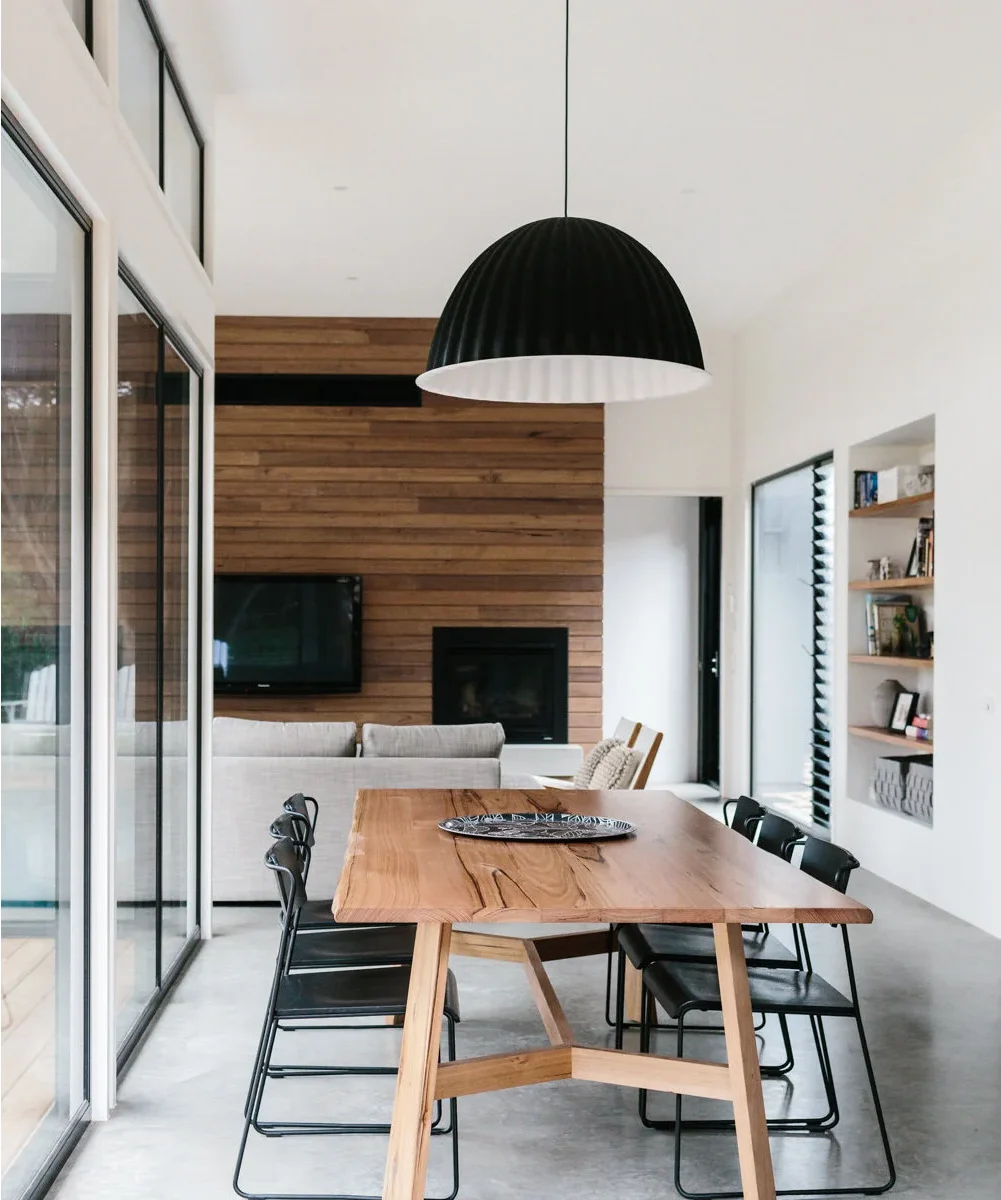 A wood dining table and black chairs run beside tall glazing, with a pale sofa and a television on a timber wall at the far end.