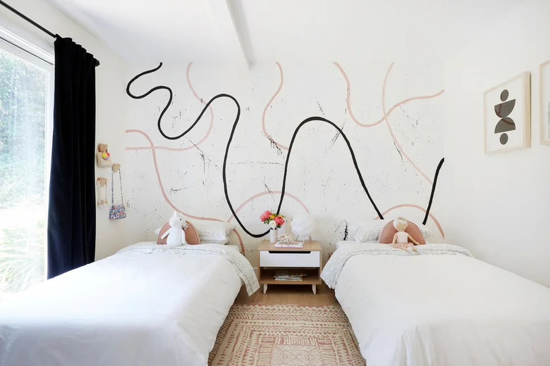 Two beds with white bedding face a white wall marked by loose black and pink lines, with a small shared bedside unit and pale patterned rug