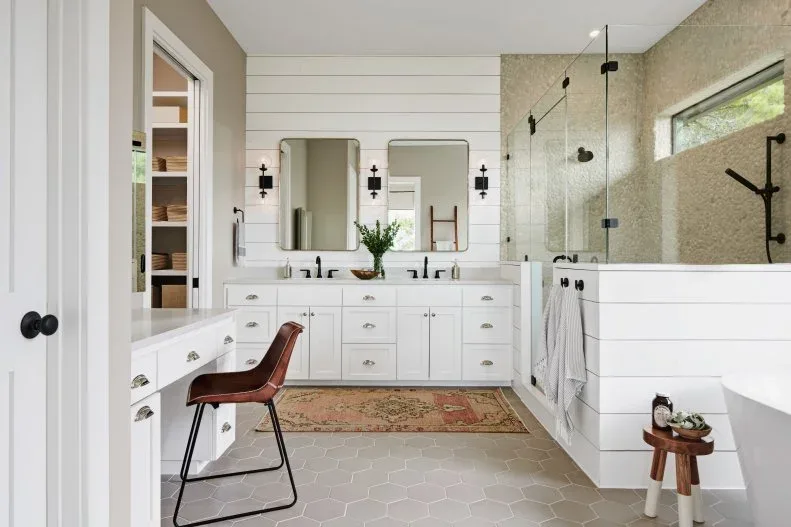 White horizontal wall panels and framed vanity cupboards sit beside a clear glass shower with dark fittings, pale hexagonal flooring and a small wood stool