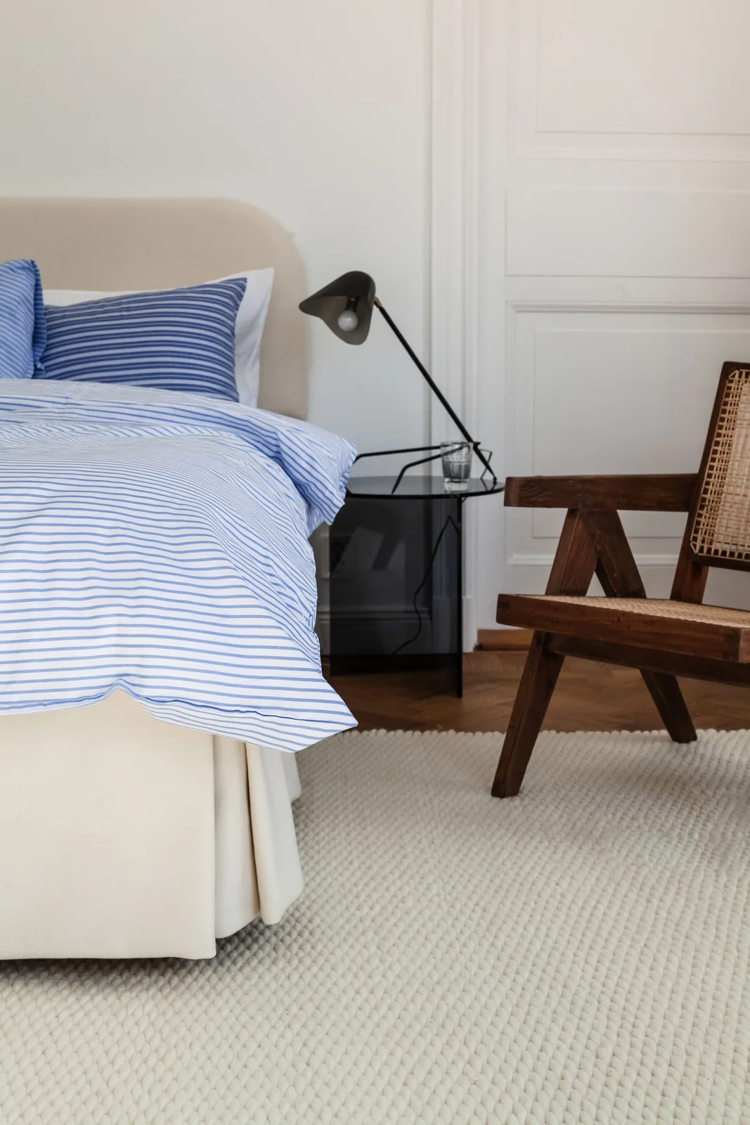 A bed with blue-and-white striped bedding sits beside a small dark glass-fronted table, a black angled lamp, a wood chair and a pale textured rug.