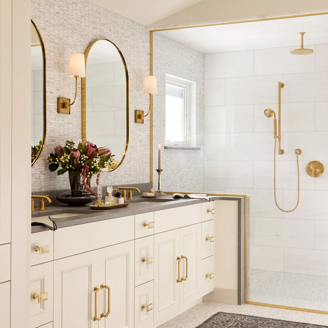 Narrow pale rectangular mosaics cover a vanity wall beneath brass-colored mirrors and lights.