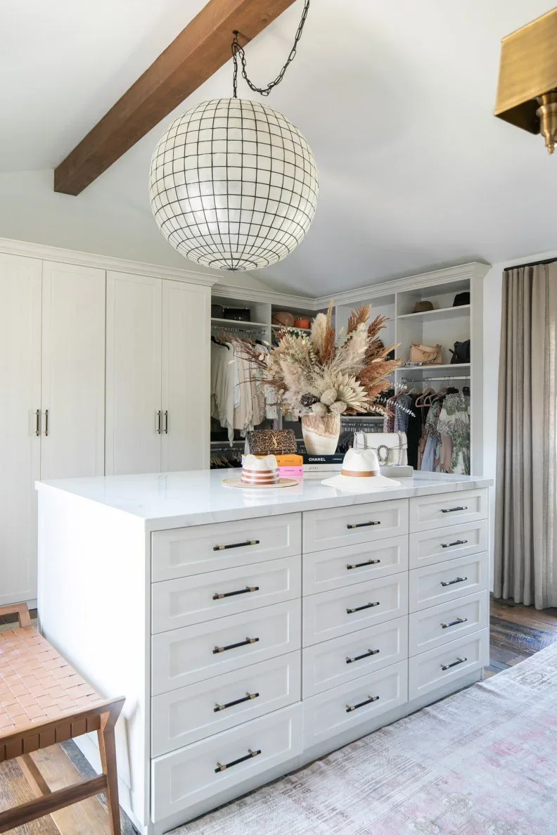A white closet island has banks of shallow drawers beneath a large globe pendant and timber ceiling beam.