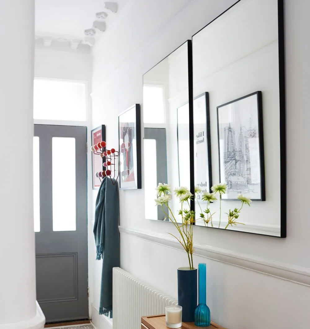 Narrow pale hallway with a large rectangular mirror above a radiator and blue vases on a small table.