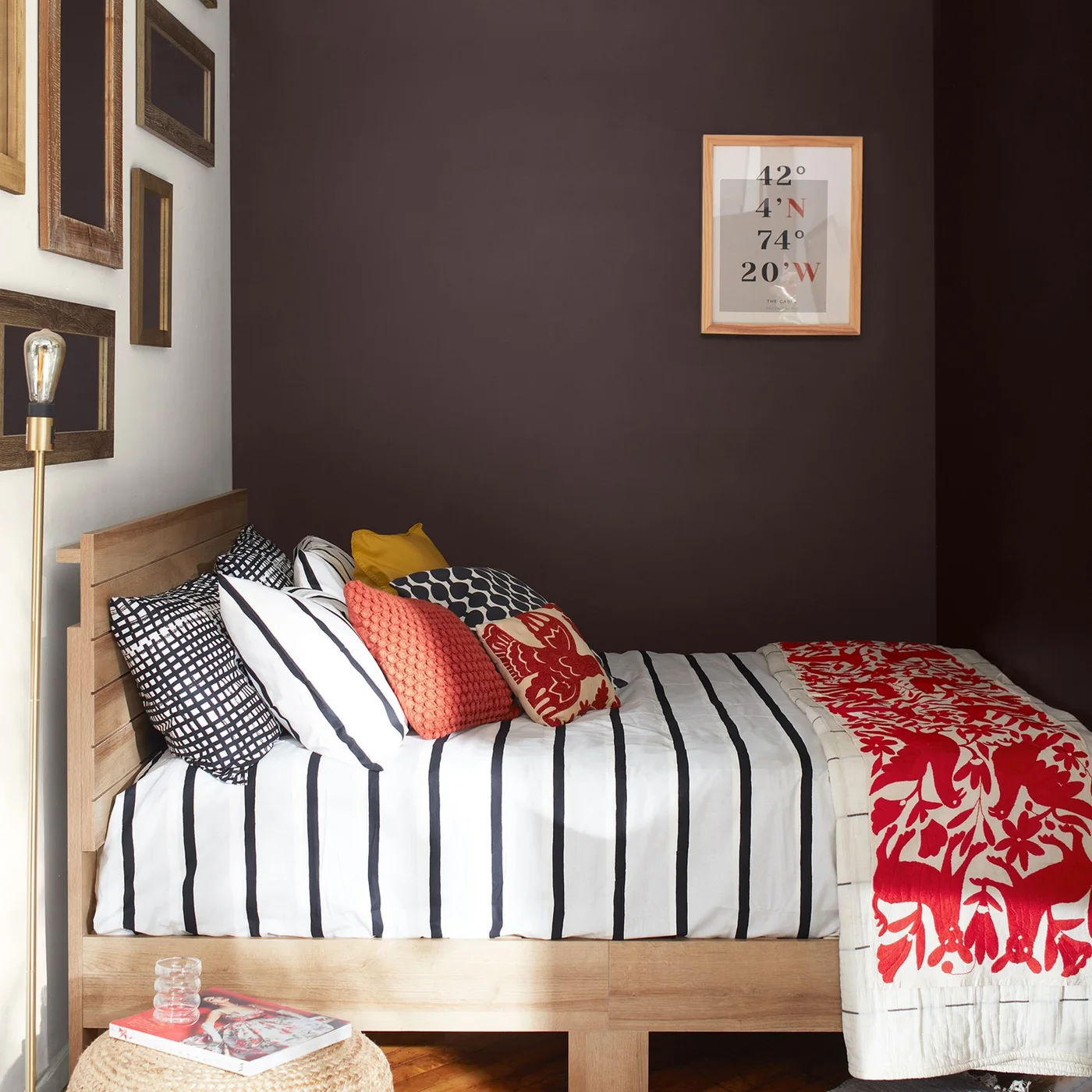 A wood bed is set lengthwise against a dark plum wall with striped bedding, red patterned textiles, a floor lamp and a small round woven table.