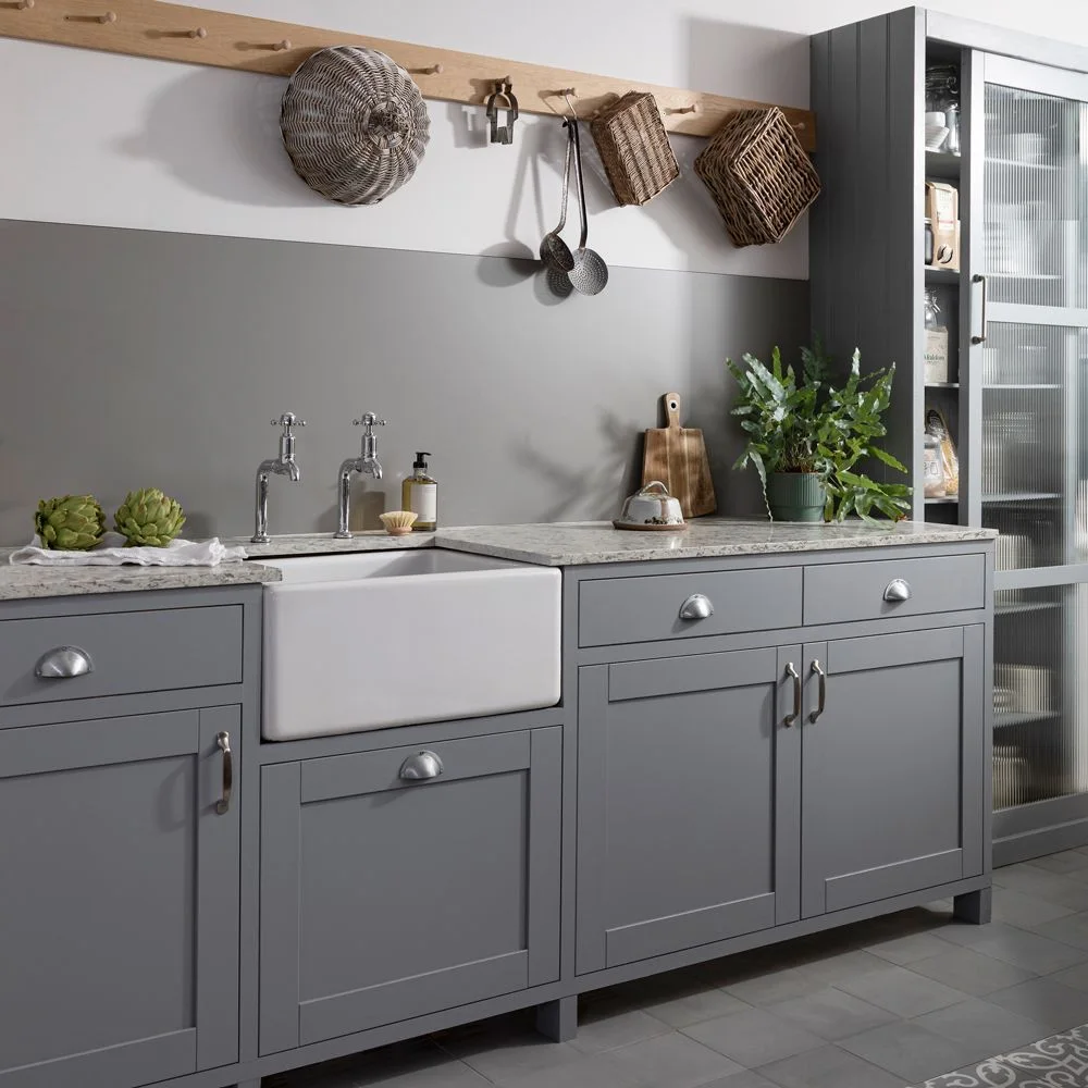 Gray paneled cupboards and drawers frame a white apron-front sink beneath a gray backsplash beside a tall ribbed-glass cabinet