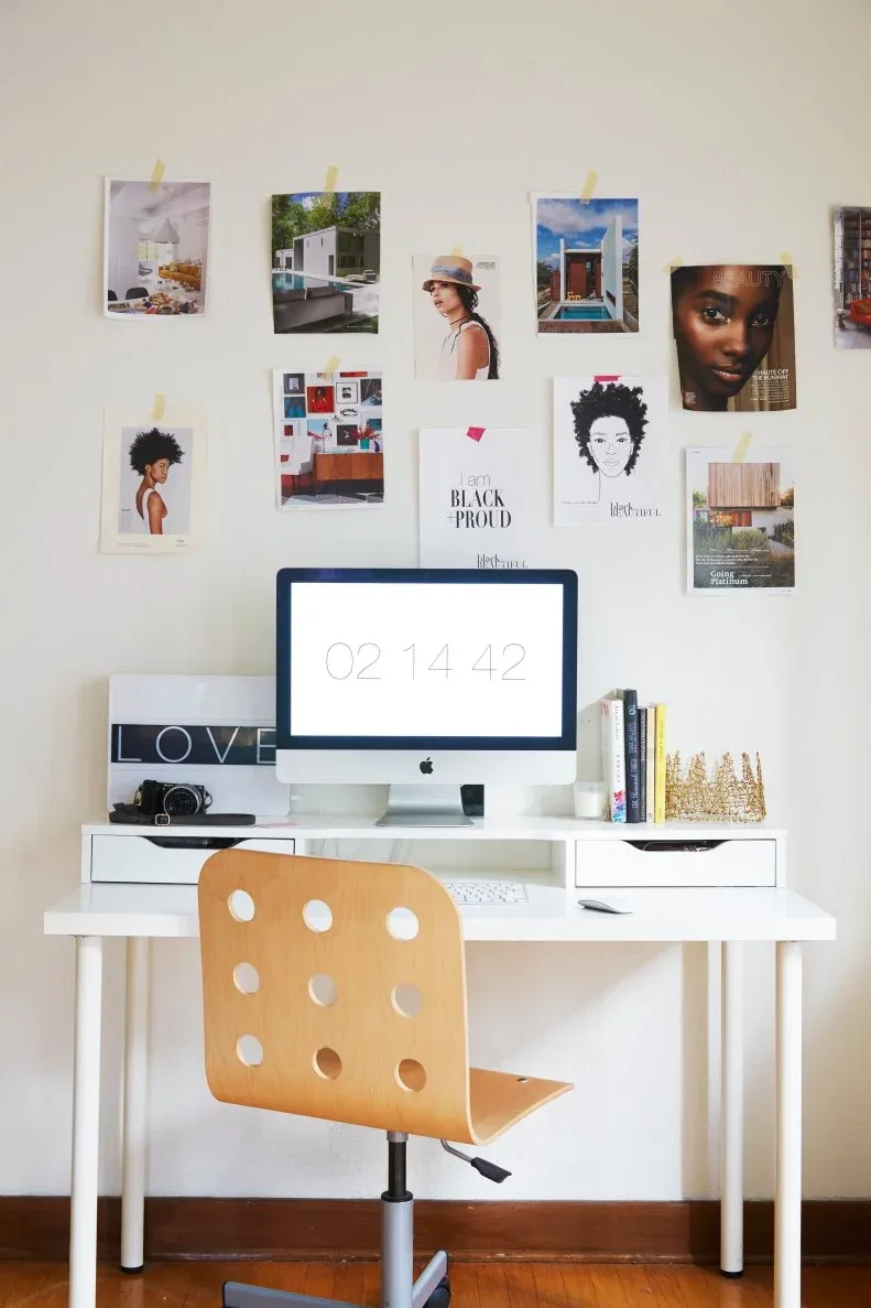 A white drawer desk with a computer and a wood swivel chair stands beneath a loose wall display of photographs, illustrations and magazine pages.