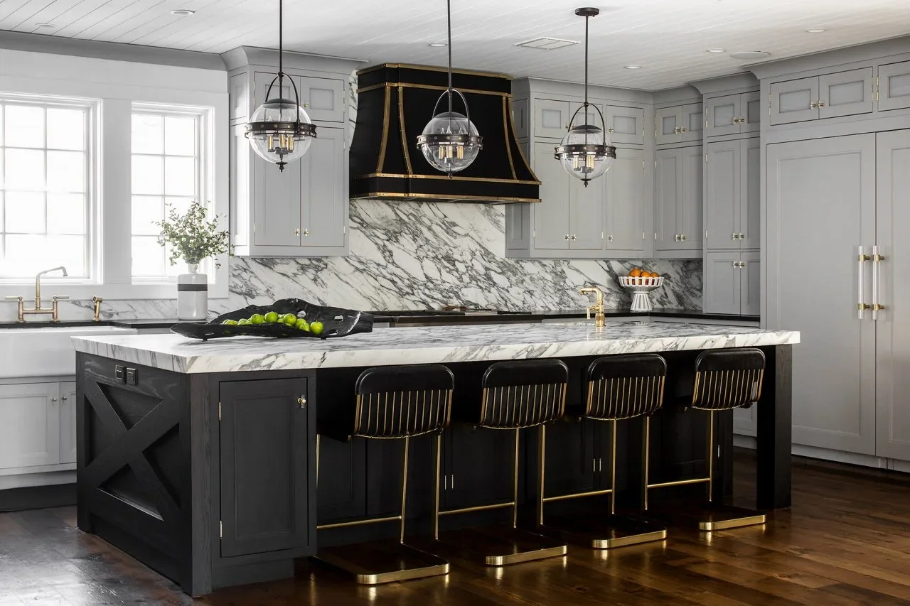 A black island and black hood with gold-colored trim contrast with gray perimeter cupboards, pale veined surfaces and four dark stools.