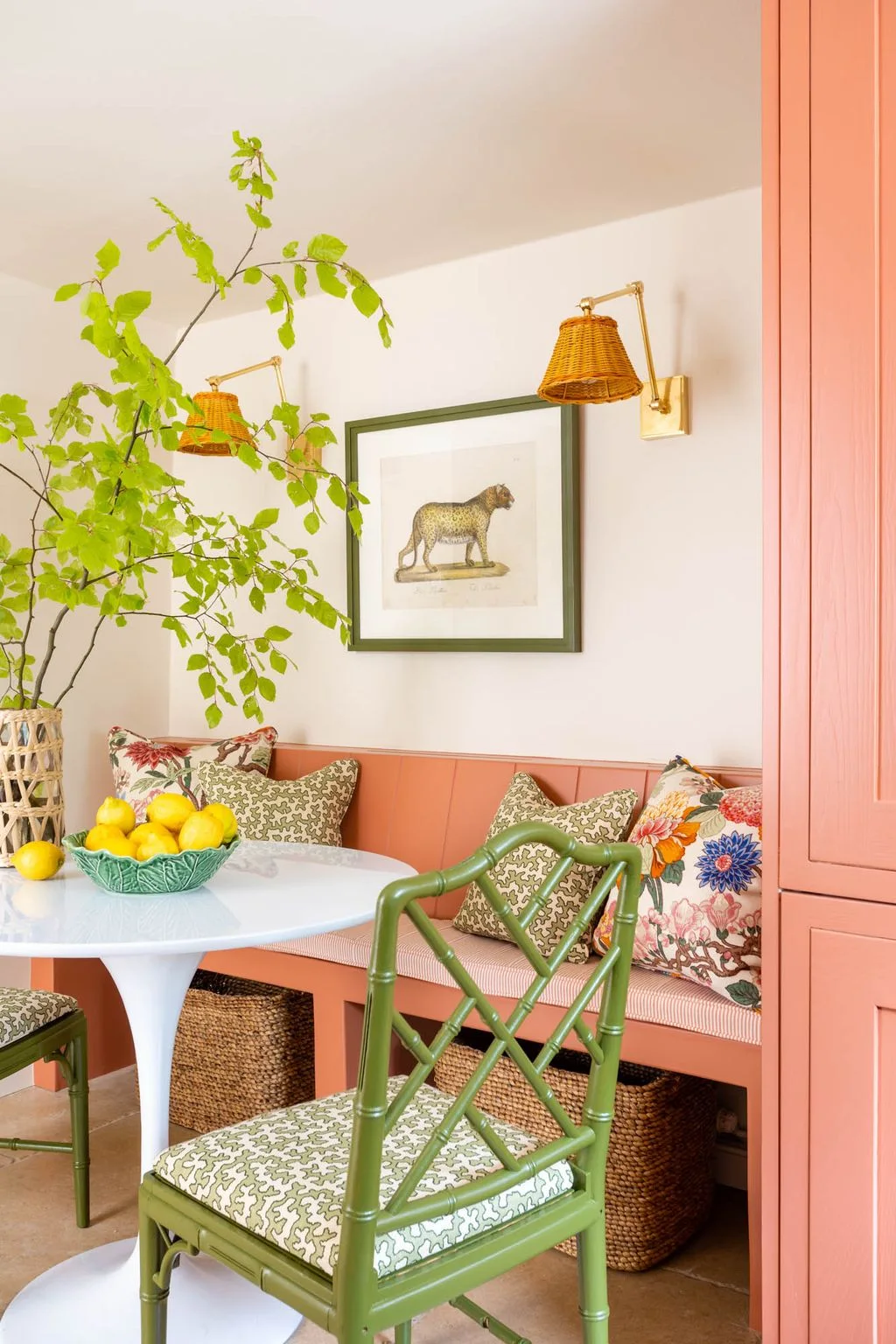 A coral built-in kitchen banquette and green chairs surround a round pale table with mixed patterned cushions and woven baskets below.