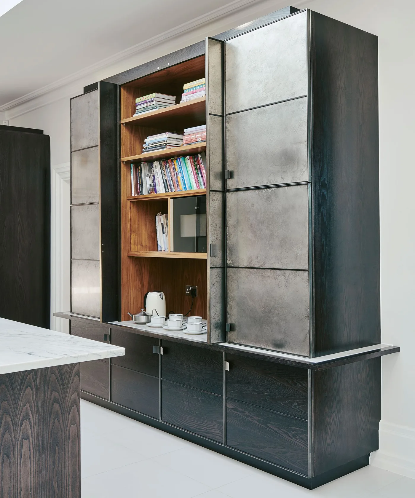 Tall kitchen storage with dark wood frame and drawers, gray metallic-looking doors, warm wood open niche, books, microwave, kettle, and cups.