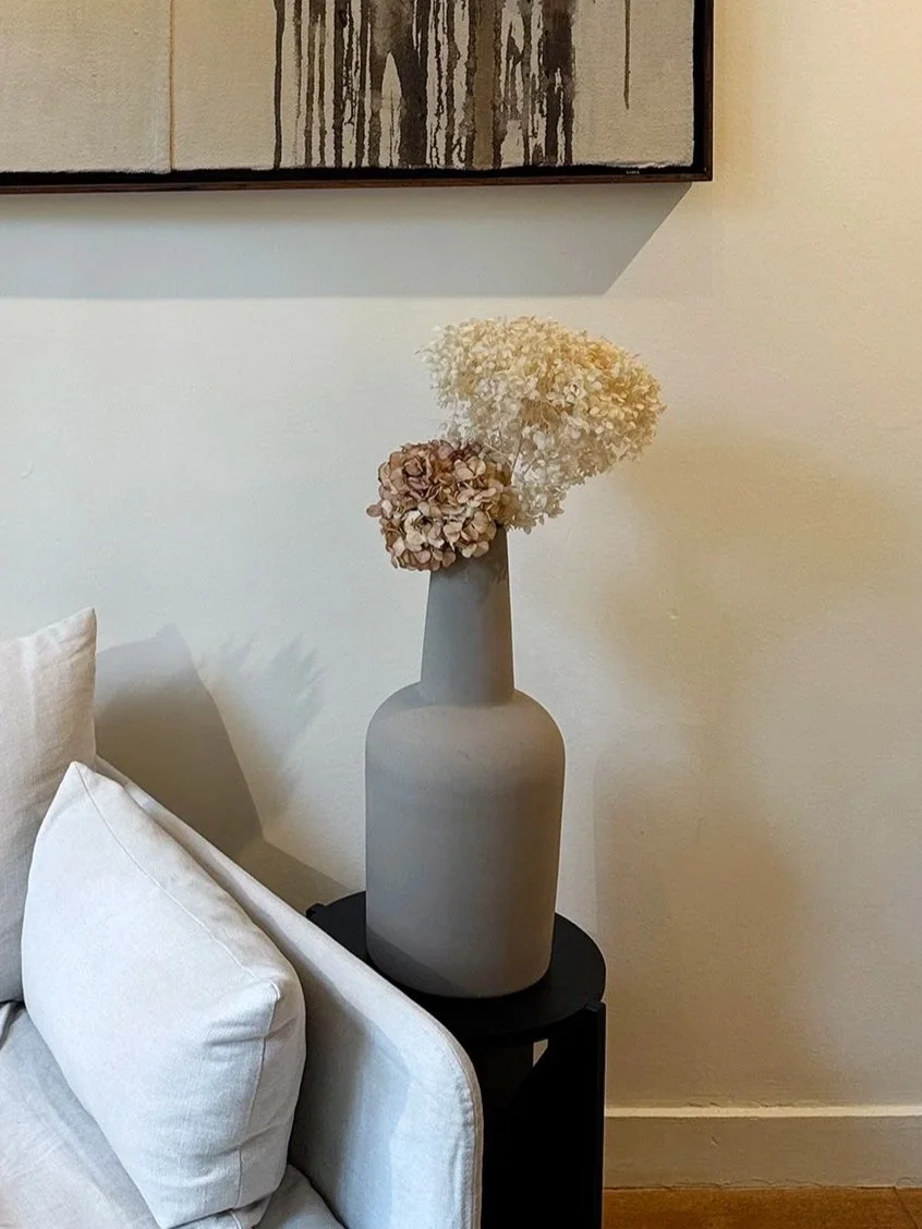 Two pale dried hydrangea heads stand in a tall gray vase on a small black side table beside a pale sofa.