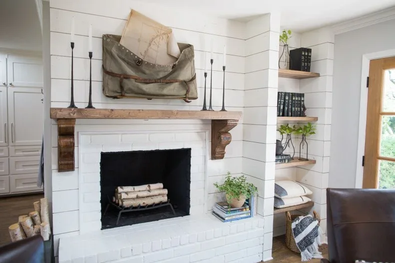 A white shiplap chimney breast has a wood mantel on large corbels, white brick around the opening and adjacent wood shelves above a small seat.